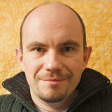 Portrait of Constantin, smiling, with a short beard, wearing a dark textured zip-up sweater against a mottled yellow background.