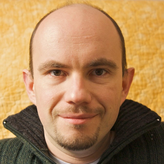 Portrait of Constantin, smiling, with a short beard, wearing a dark textured zip-up sweater against a mottled yellow background.