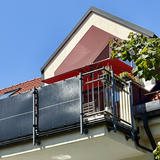 A multi-story residential building with red clay tile roofing under a clear blue sky. The upper level features a balcony with metal railings and solar panels mounted as balcony power plant system (Balkonkraftwerk). Above the balcony is a triangular dormer window with cream and reddish-brown colored walls, topped by a red awning. Green leafy vines or foliage are visible growing around the right side of the building.