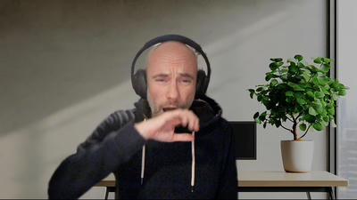 Constantin, wearing over-ear headphones and a dark hoodie, yawning during a video call, sitting at a desk with a generic, uninspiring stock video background behind him, including a monitor, and a potted green plant on the desk near a window.