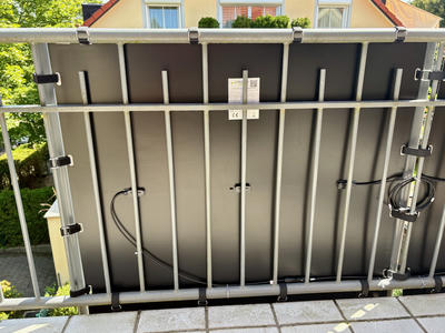 A solar panel mounted vertically on straps in a metal frame structure on a balcony. The panel has cables connecting to an adjacent panel and there are small identification labels visible. In the background, there is a yellow house with red roof tiles, green trees, and landscaping visible below the balcony.