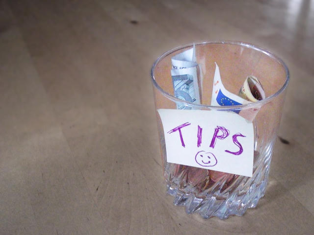 A glass jar labeled “TIPS” with a smiley face, filled with folded banknotes and coins, sitting on a wooden floor.