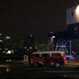 A colorful old Volkswagen van sits parked on a wet dockside street at night, with city lights and a crane visible in the background.