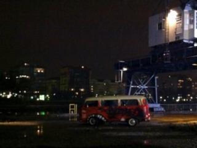 A colorful old Volkswagen van sits parked on a wet dockside street at night, with city lights and a crane visible in the background.